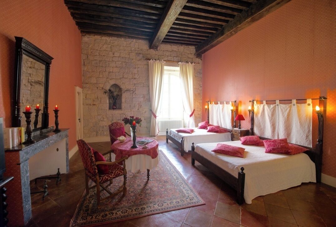 Une chambre avec deux lits, un coin salon avec un fauteuil et une table, une cheminée et un mur en pierre.