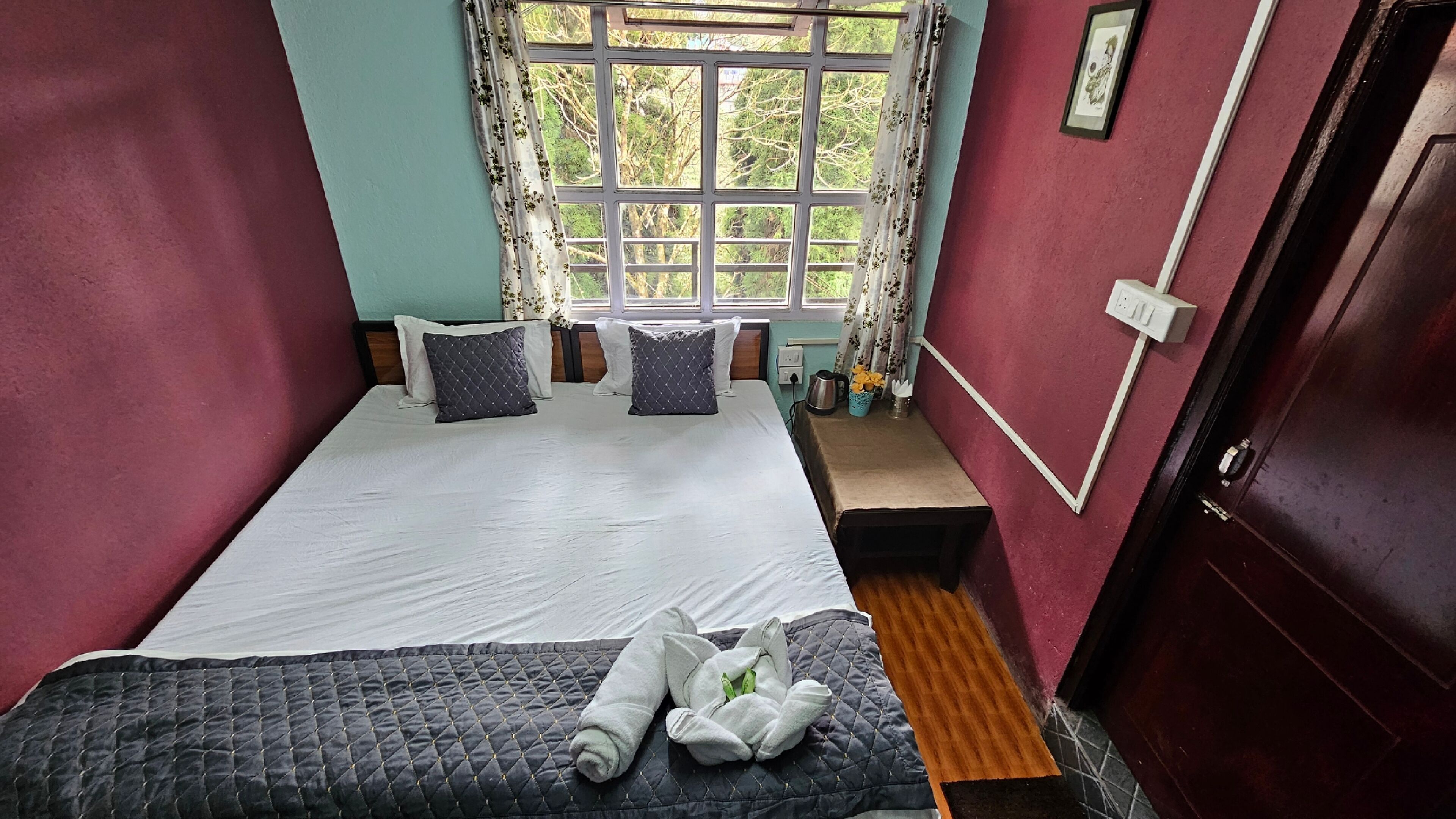 Ein ordentlich bezogenes Bett mit Kissen und einer gesteppten Decke, ein kleiner Tisch mit Blumen in einer Vase und ein Fenster mit Vorhängen.