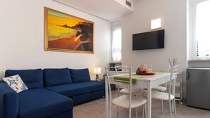 Uma sala de estar moderna com um sofá azul, uma mesa de jantar com cadeiras brancas, um quadro na parede e uma TV de tela plana instalada acima da mesa.