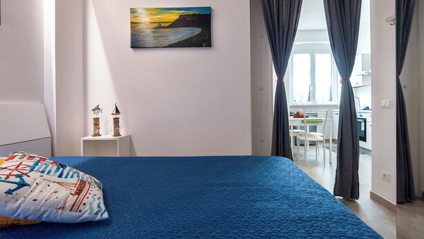 Quarto com colcha azul, pintura de pôr do sol, mesa pequena com dois objetos decorativos e vista para uma sala de jantar através de um vão.