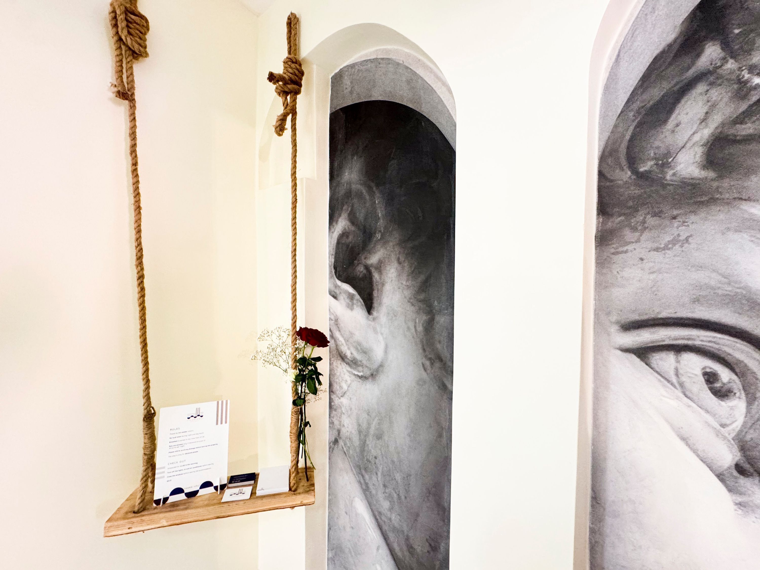 A room with a wooden swing, a black and white painting, and a small vase with a red rose.