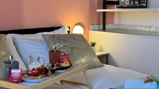 Un plateau de petit-déjeuner bien présenté, avec une théière, un pichet et une assiette de fruits, posé sur un lit agrémenté d’oreillers et d’une table de chevet.