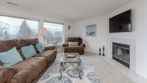 A living room with a brown leather sofa, a glass coffee table, a fireplace, and a large TV on the wall. - Park Pointe B102 – Walk to the Beach & Enjoy the Pool & Hot Tub! (Chelan)