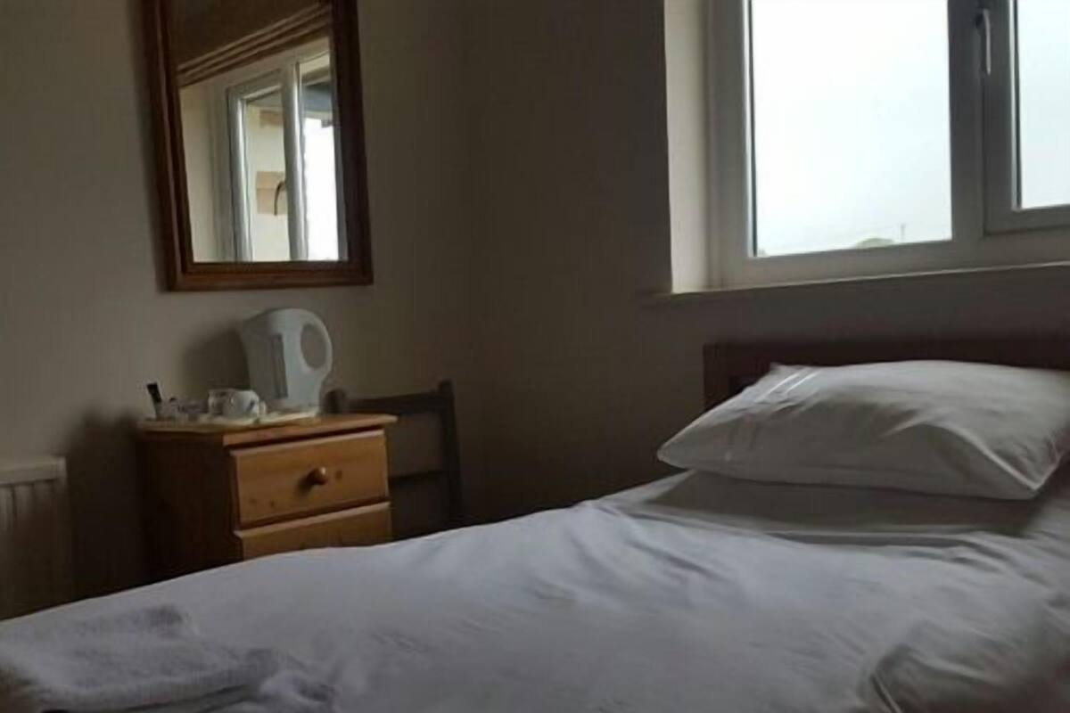A neatly made bed with white linens, a bedside table with a kettle, and a chair.