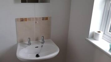 Une salle de bain avec un lavabo blanc, deux robinets, un miroir au-dessus du lavabo et une fenêtre sur le rebord de laquelle repose un rouleau de papier toilette.