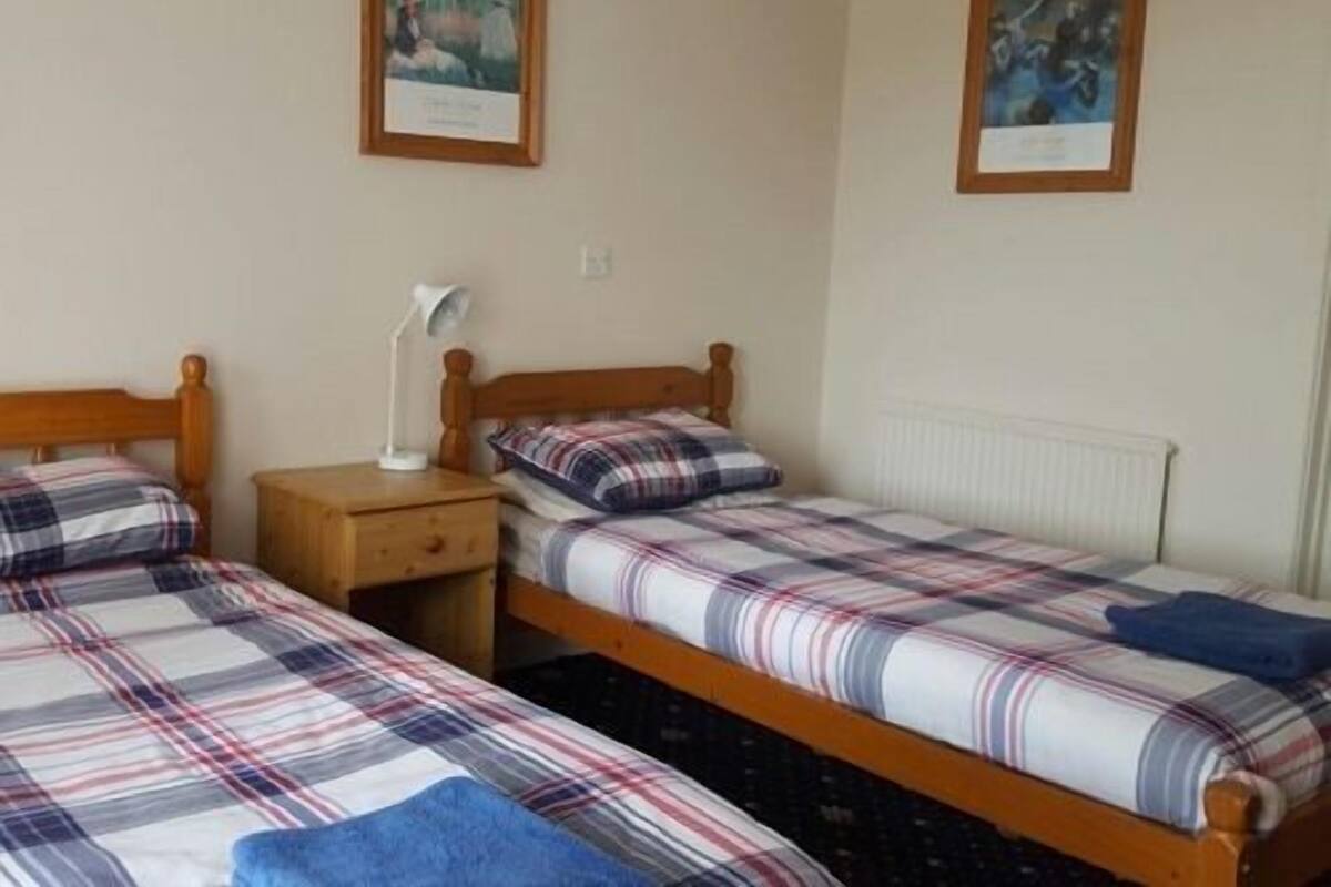 A room with two single beds, each with a plaid bedspread and matching pillows. There is a nightstand with a lamp between the beds.