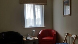 A room with a window, a red armchair, a wooden chair, and a small table with a lamp and a box. - A35 PitStop (Axminster)