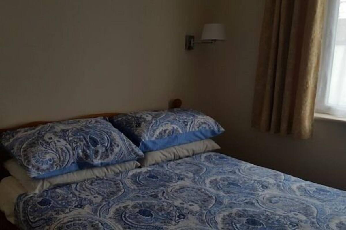 A bed with a blue patterned bedspread and pillows, a wall-mounted lamp, and a window with curtains.
