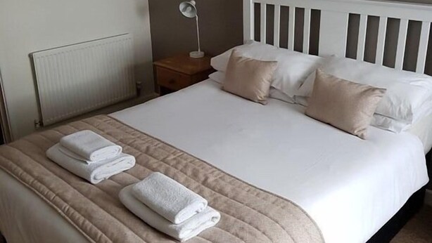 A neatly made bed with white linens and beige pillows, a bedside lamp, and a framed picture on the wall. - A35 PitStop (Axminster)