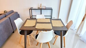 A dining area with a table set for four, surrounded by white chairs with wooden legs. A grey sofa is positioned to the left, and a small black table with a Buddha statue and a sign is against the wall.