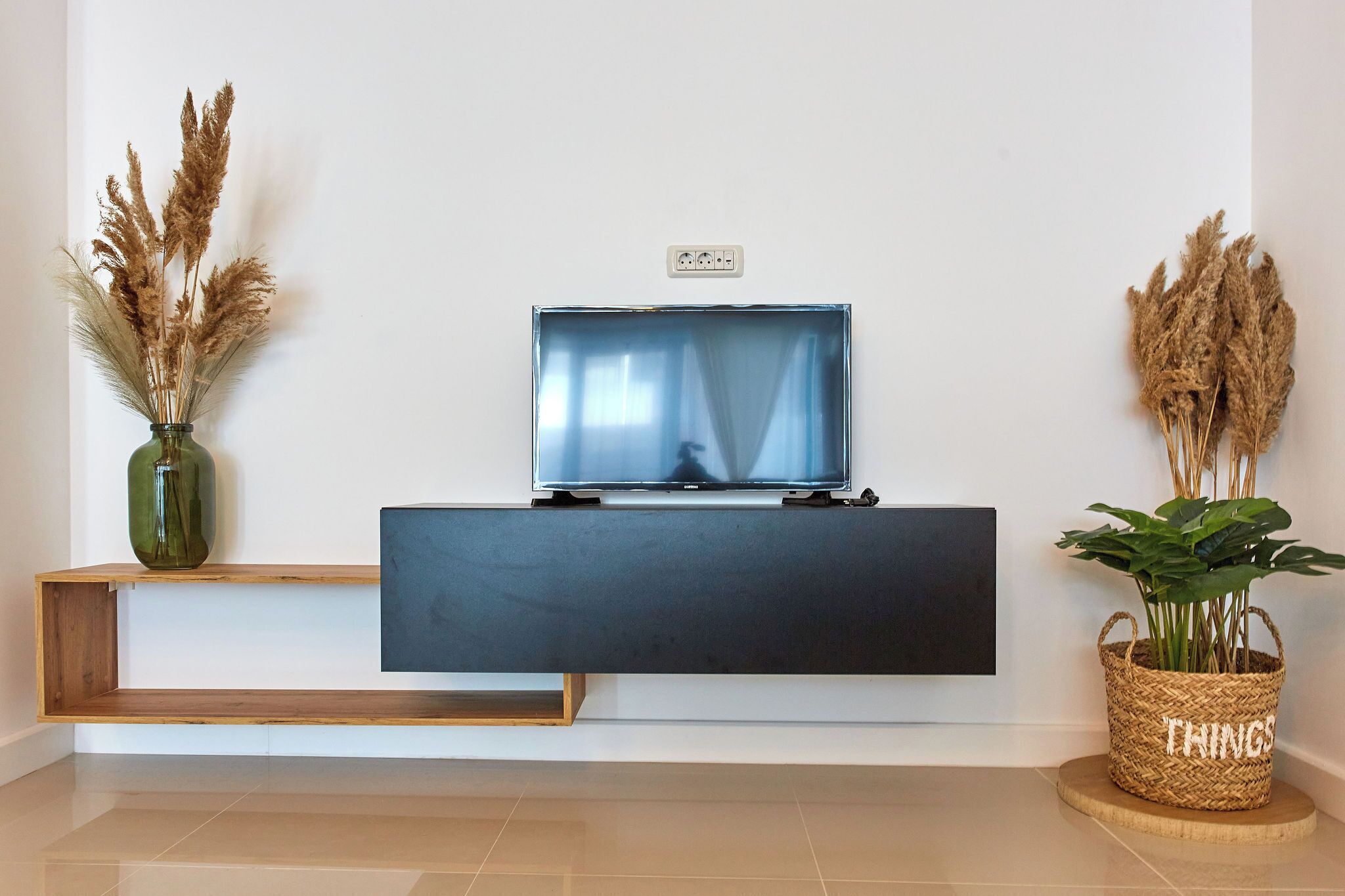A modern living room with a flat-screen TV on a black stand, a wooden bench, and two decorative plants.