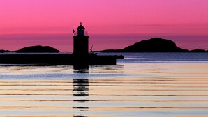 Silueta d'un far davant un cel rosat i morat, aigua tranquil·la i terres llunyanes.