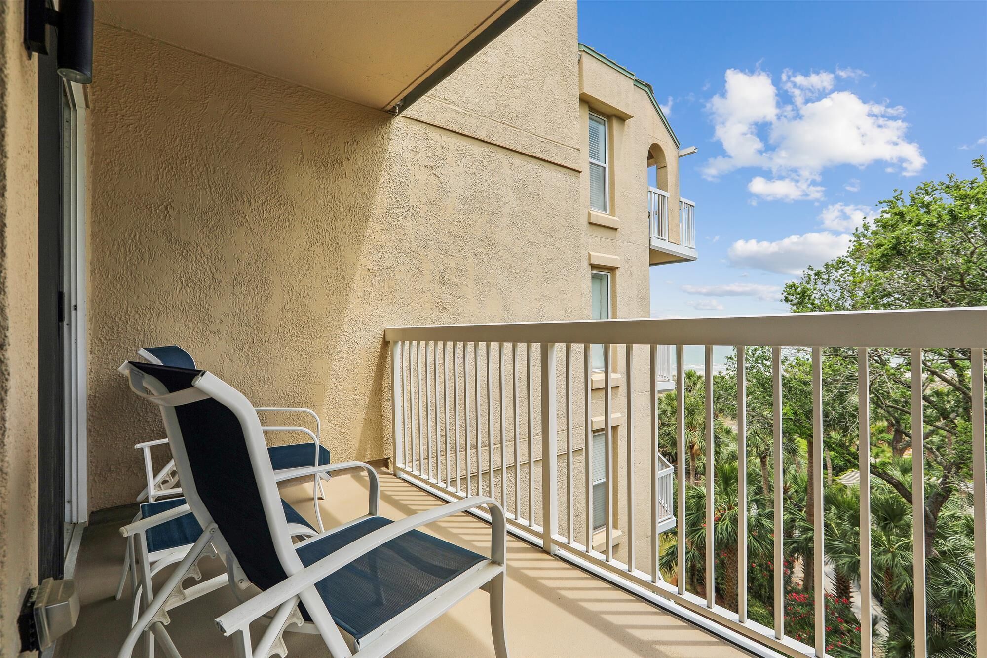 A balcony with two lounge chairs, a white railing, and a view of a building and trees.
