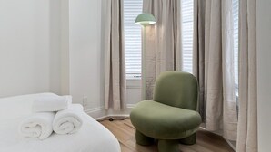 Ein Hotelzimmer mit einem Bett, aufgerollten weißen Handtüchern, einem grünen Sessel, einer Stehlampe und beigen Vorhängen.