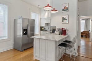 Une cuisine moderne dotée d’un réfrigérateur en acier inoxydable, de meubles blancs, d’un îlot central avec un comptoir en marbre et de luminaires suspendus rouges.