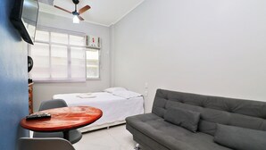 A small, sparsely furnished room with a grey sofa, a small round table, a bed with white bedding, a ceiling fan, and a window with blinds. - Santa Clara Studio (Rio de Janeiro)