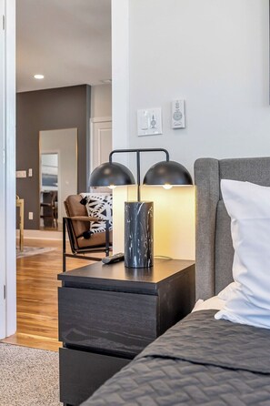 Moderni hotellihuone, jossa on sänky, yöpöytä lampun kanssa, tuoli ja näkymä käytävälle.