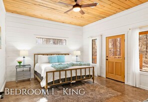 A bedroom with a wooden ceiling fan, a bed with a headboard, two bedside tables with lamps, a window with curtains, and a door with a view of trees.