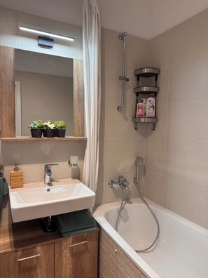 A modern bathroom with a white sink, wooden cabinet, and a bathtub. There are potted plants on a shelf above the sink and a showerhead mounted on the wall. - Apart 555 New & Fully Equipped with Garage & 15 Min to City Center (Bratislava)