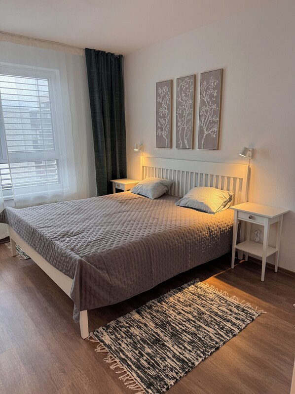 A bedroom with a bed, two bedside tables, a nightstand, a window with curtains, and wall art. - Apart 555 New & Fully Equipped with Garage & 15 Min to City Center (Bratislava)