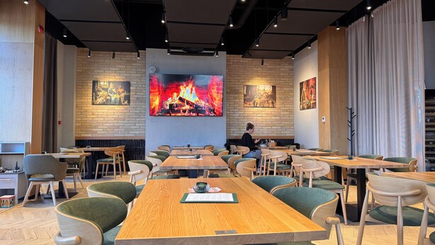 Een modern restaurant met houten tafels en groene stoelen met kussens, bakstenen muren en grote schilderijen van een vuur.