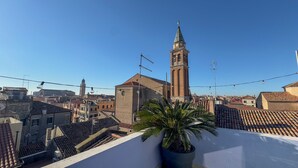 Une vue sur un paysage urbain avec un clocher d’église imposant, des toits en tuiles de couleur terre cuite et un pot de fleurs sur un balcon.