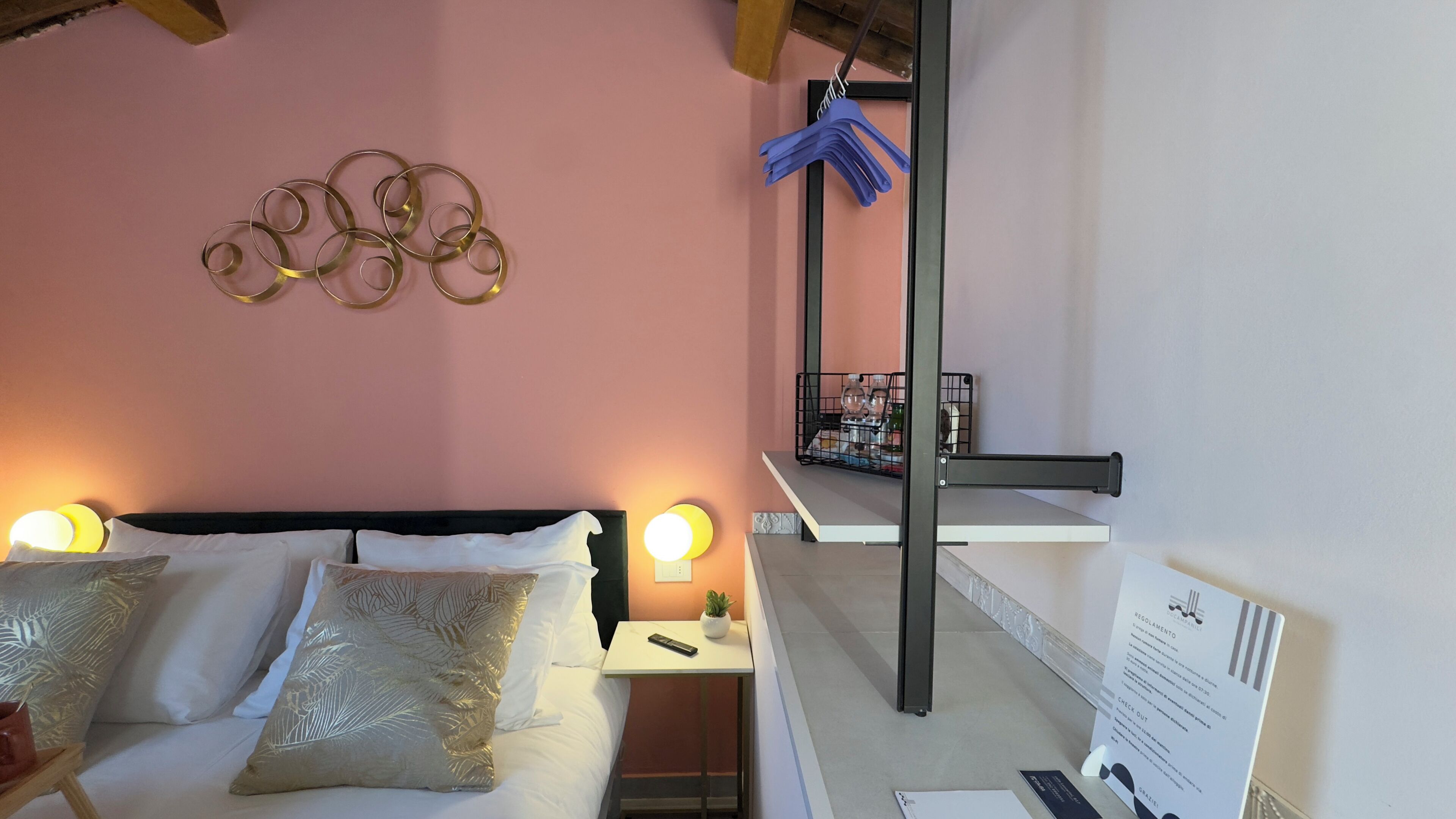 Habitación de hotel con cama, lámparas de noche, aplique de pared, una mesita con una planta y una estantería con objetos.