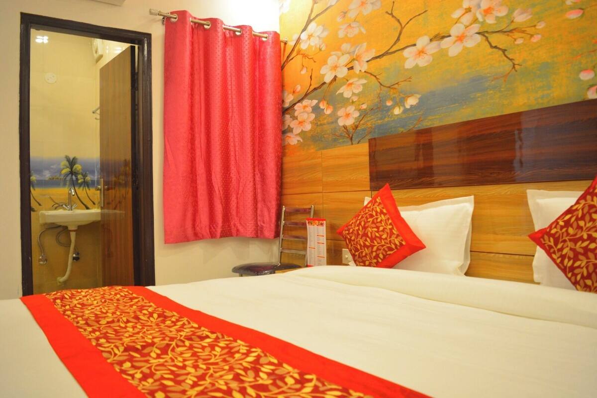 A hotel room with a bed, a wooden headboard, a red curtain, and a wall painting of cherry blossoms.