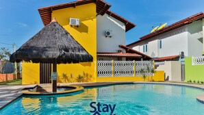 A swimming pool with a 'Stay Hall' sign, surrounded by buildings with yellow and white walls, a thatched roof structure, and a white fence. - Casa 11 Villa Giardino-SH033 (Tamandaré)