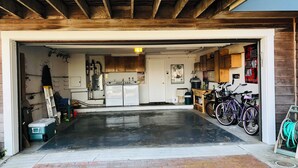 A garage with a black floor, wooden beams, a refrigerator, a washing machine, and bicycles.