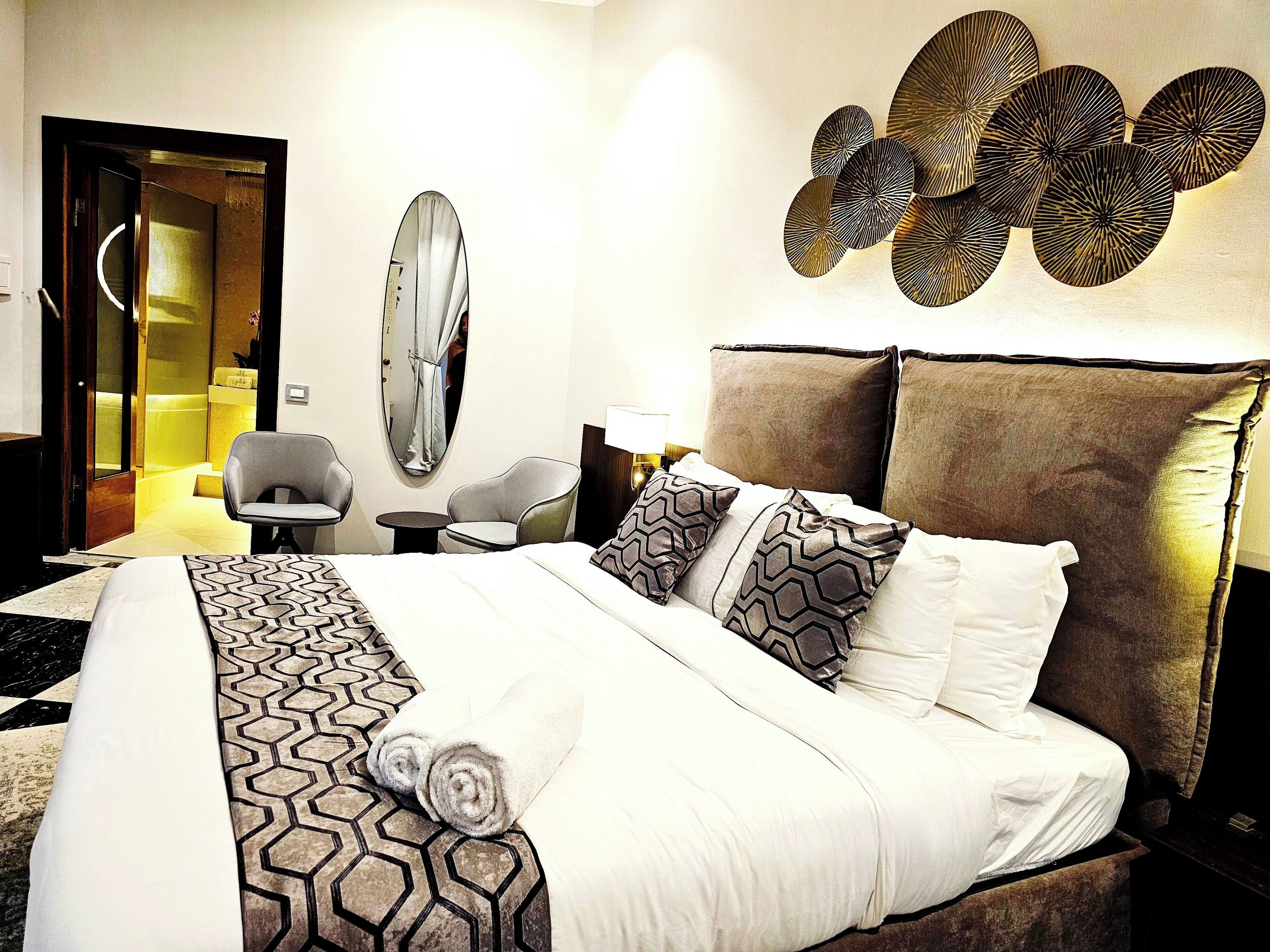 A hotel room with a large bed, a round mirror, a wall art piece, and a seating area with a small table and chairs.