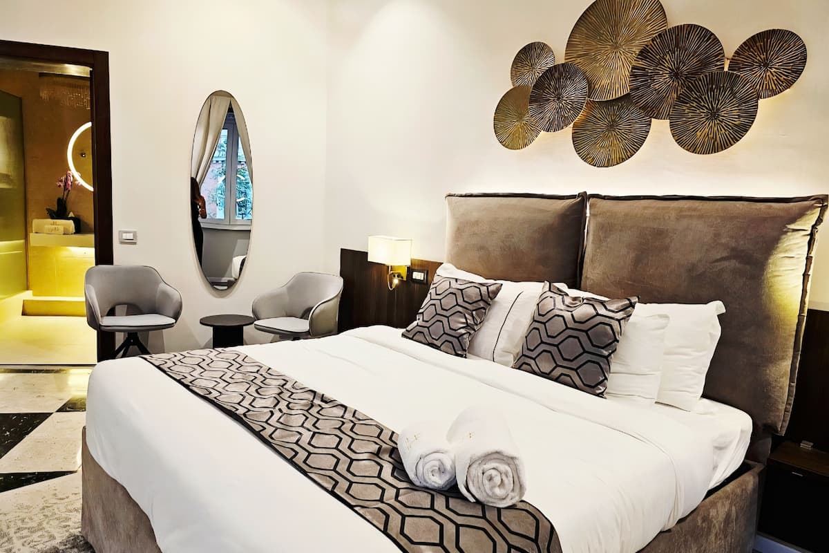 A modern hotel room with a large bed, two armchairs, a small table, a mirror, and a decorative wall piece.