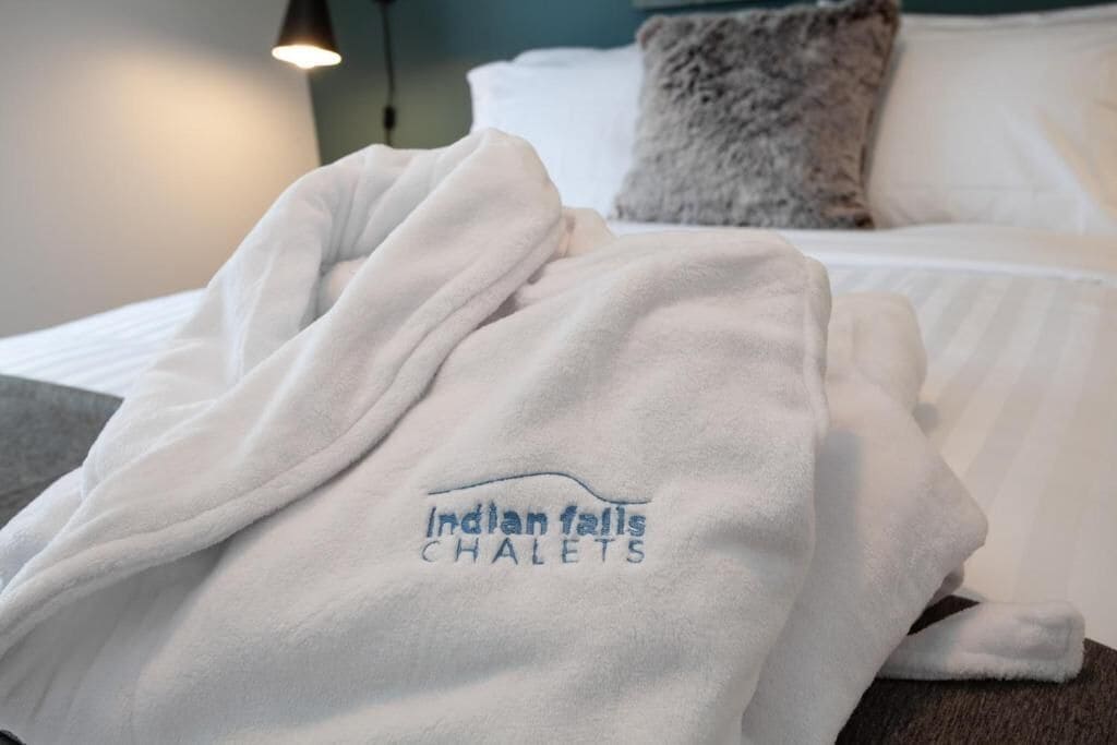 Ein Hotelzimmer mit einem ordentlich bezogenen Bett, einem flauschigen Kopfkissen und einem Bademantel mit der Aufschrift "Indian Falls Chalets".