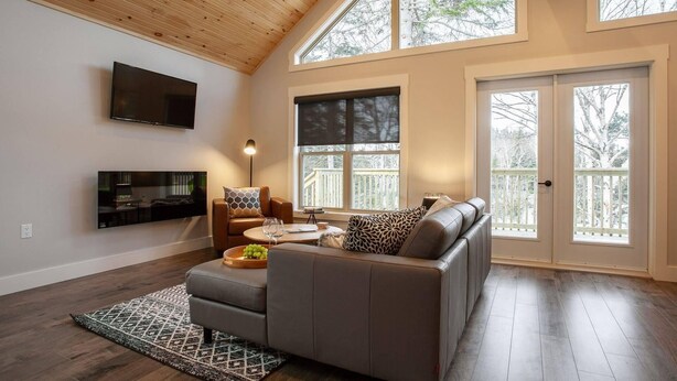 A spacious living room with a high ceiling, a large window, a flat-screen TV, a sofa, and a coffee table. - Indian Falls Chalets  (Springdale)