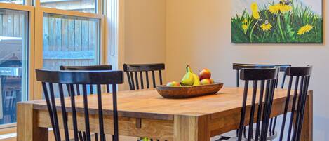 Uma área de refeições com mesa e cadeiras de madeira, uma fruteira e um quadro na parede.