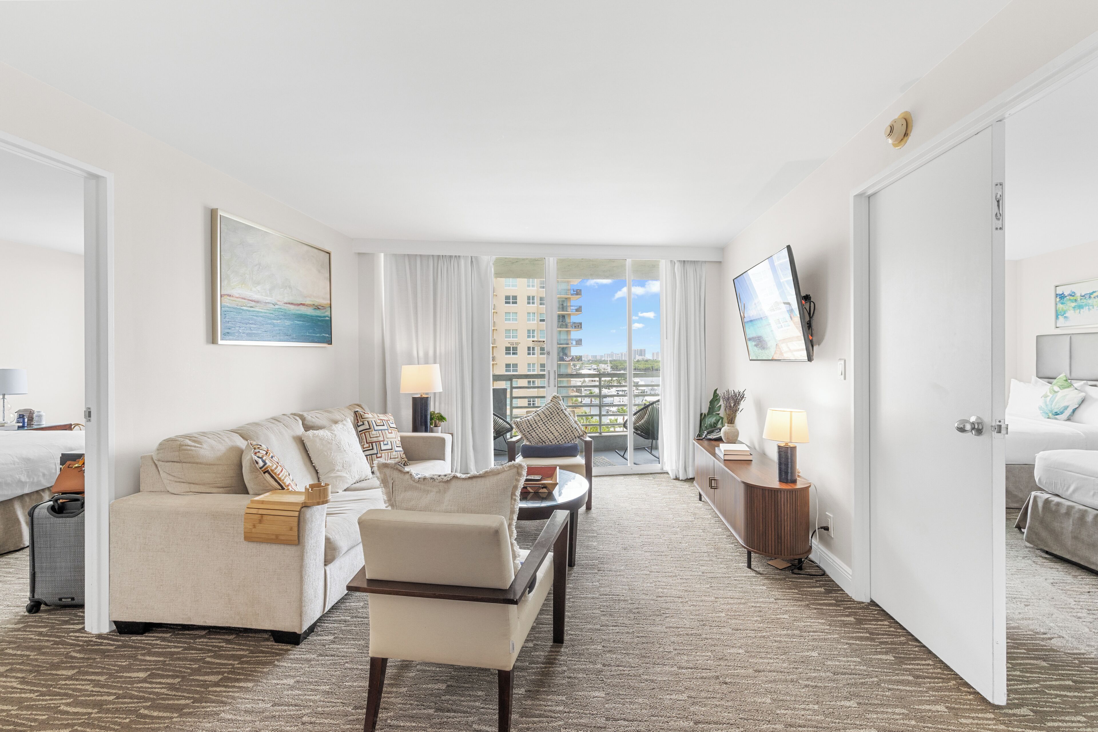A hotel room with a sofa, armchair, a dining table, a TV mounted on the wall, and a view of the city through the window.