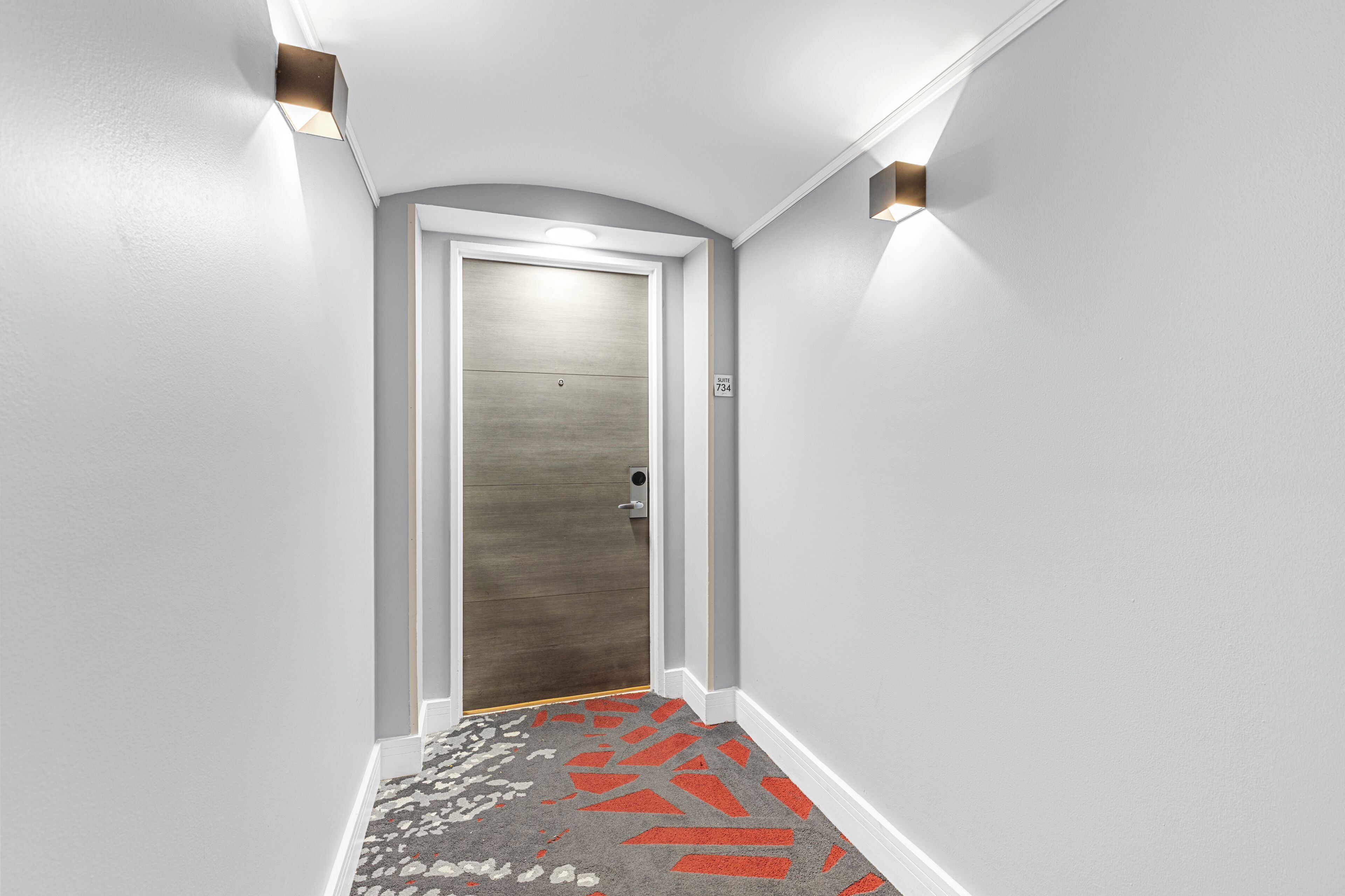 Un couloir avec une porte, un tapis aux motifs rouges et gris, et deux lampes fixées au mur.