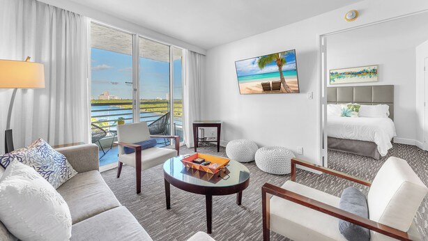 A hotel room with a large window, a sofa, a coffee table, a bed, and a painting of a beach scene. - Eternal Sunrise (Fort Lauderdale)