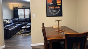 A living room with a black leather couch, a wooden dining table with chairs, and a wall-mounted poster of West Virginia's statehood.