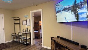 A living room with a TV mounted on the wall, a wooden console table, a black metal shelf unit, and a bunk bed in the background.