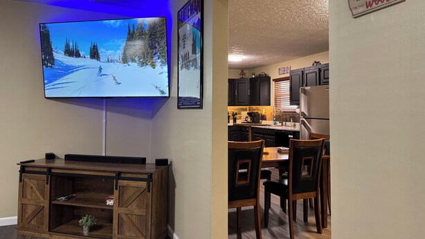 A living room with a mounted TV, wooden entertainment unit, and a dining area with a kitchen in the background.