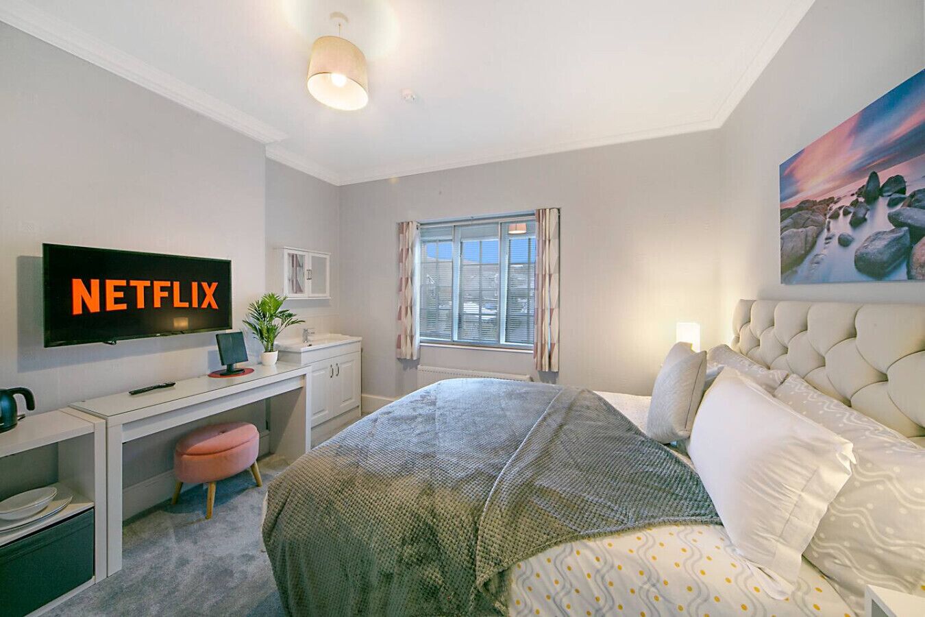 Een moderne hotelkamer met een bed, een bureau met een computer, een tv waarop 'NETFLIX' staat, een raam met gordijnen en een schilderij aan de muur.