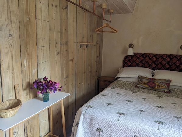 A bedroom with a wooden wall, a bed with a patterned headboard, a bedside table with a basket, and a vase with flowers. - Charming cottage on the River Boyne,Fishing ,Kayaking,World Heritage sites (Slane)