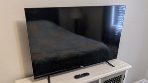 Un televisor sobre un mueble blanco con frente de malla, cajones y control remoto.