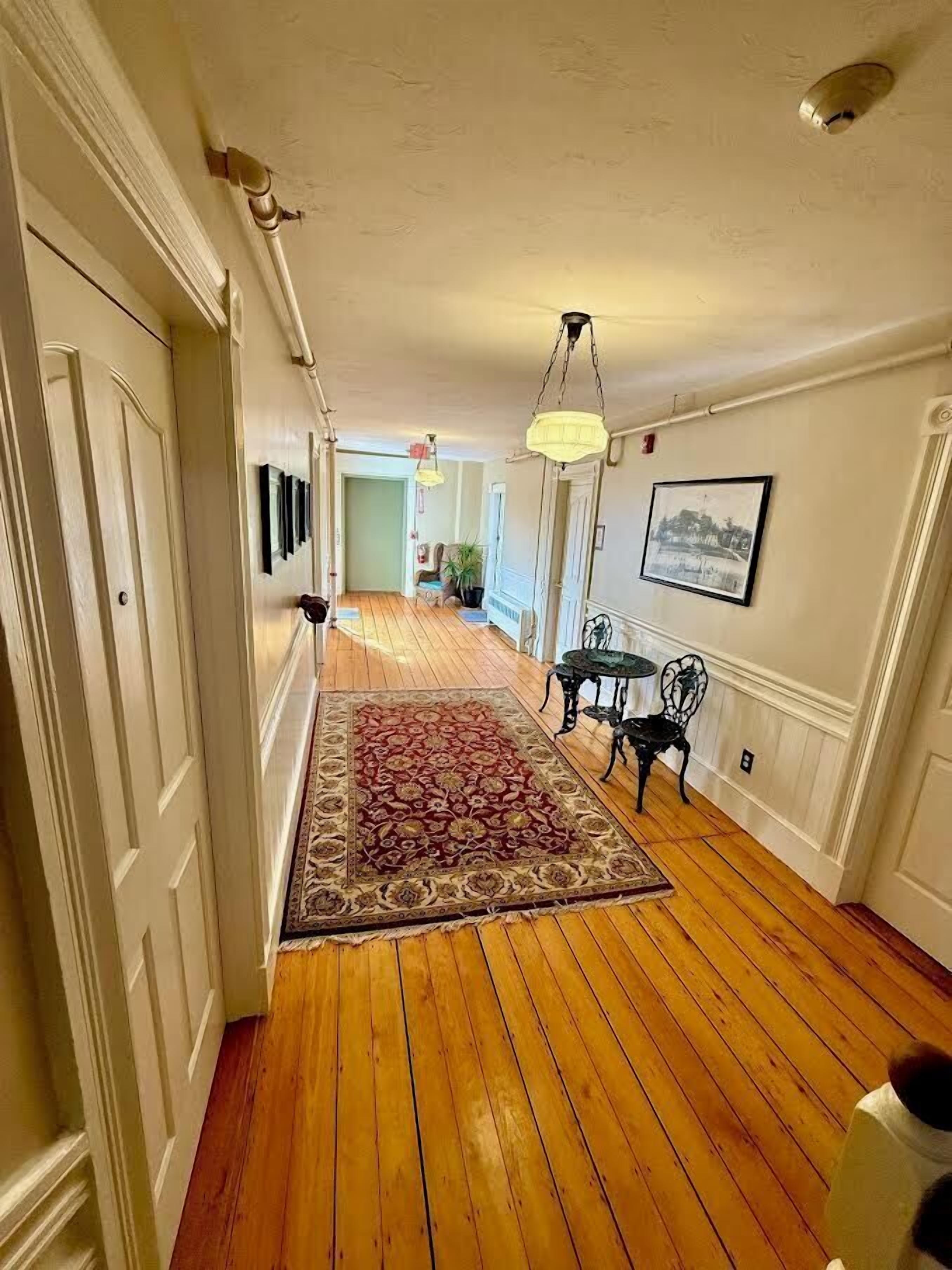 A hallway with wooden flooring, a patterned rug, and a table with chairs.