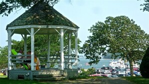 Un gazebo con tetto in tegole e pareti bianche, circondato da erba e alberi, con vista su un corpo d'acqua e imbarcazioni.