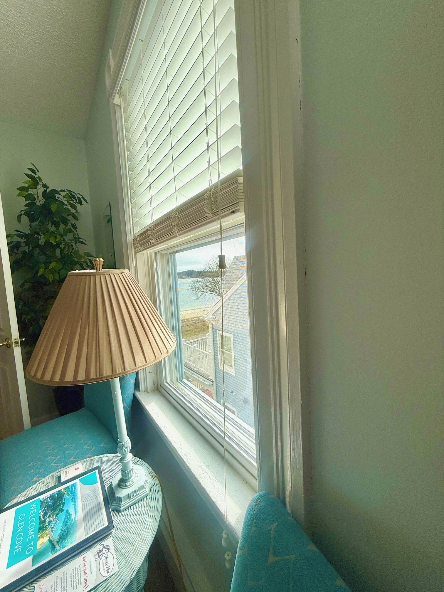 A corner room with a window, a lamp, a small table with a brochure, and a blue chair.