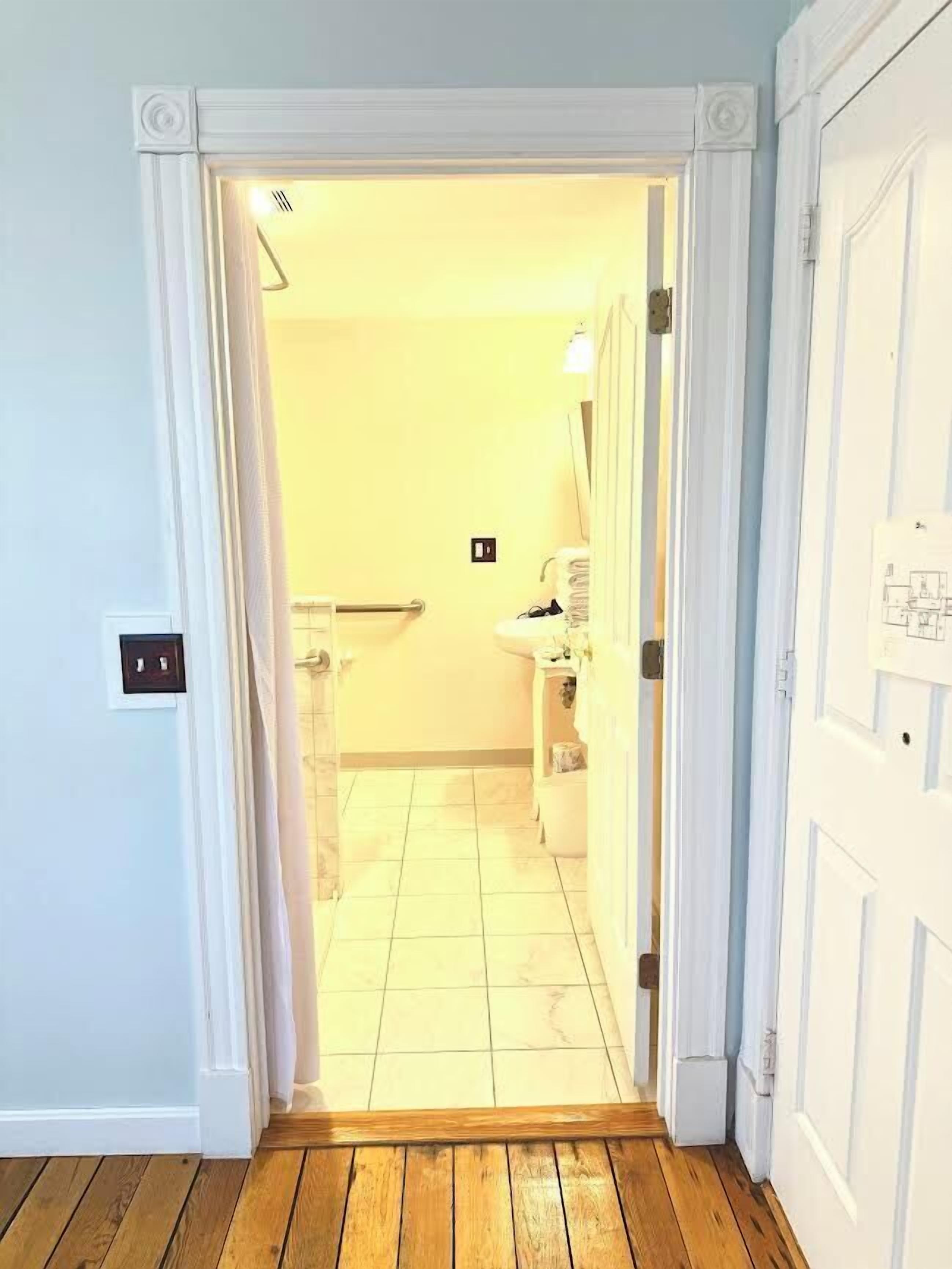 Un couloir menant à une salle de bain carrelée, équipée d’un lavabo et de toilettes.