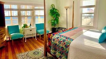 A bedroom with a four-poster bed, a desk with a chair, a potted plant, and a view of the outdoors through large windows.