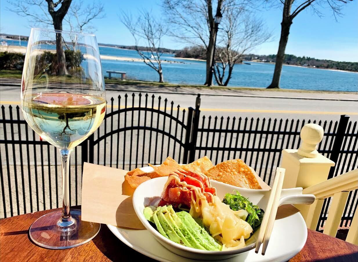 A table with a glass of white wine, a plate of food including vegetables and bread, and a view of a body of water and trees in the background.